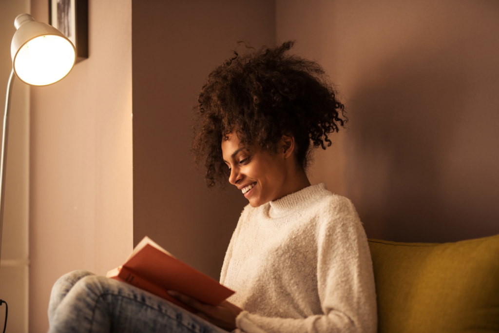 Schwarz gelesene Person sitzt lächelnd mit einem Buch auf dem Schoß und liest.